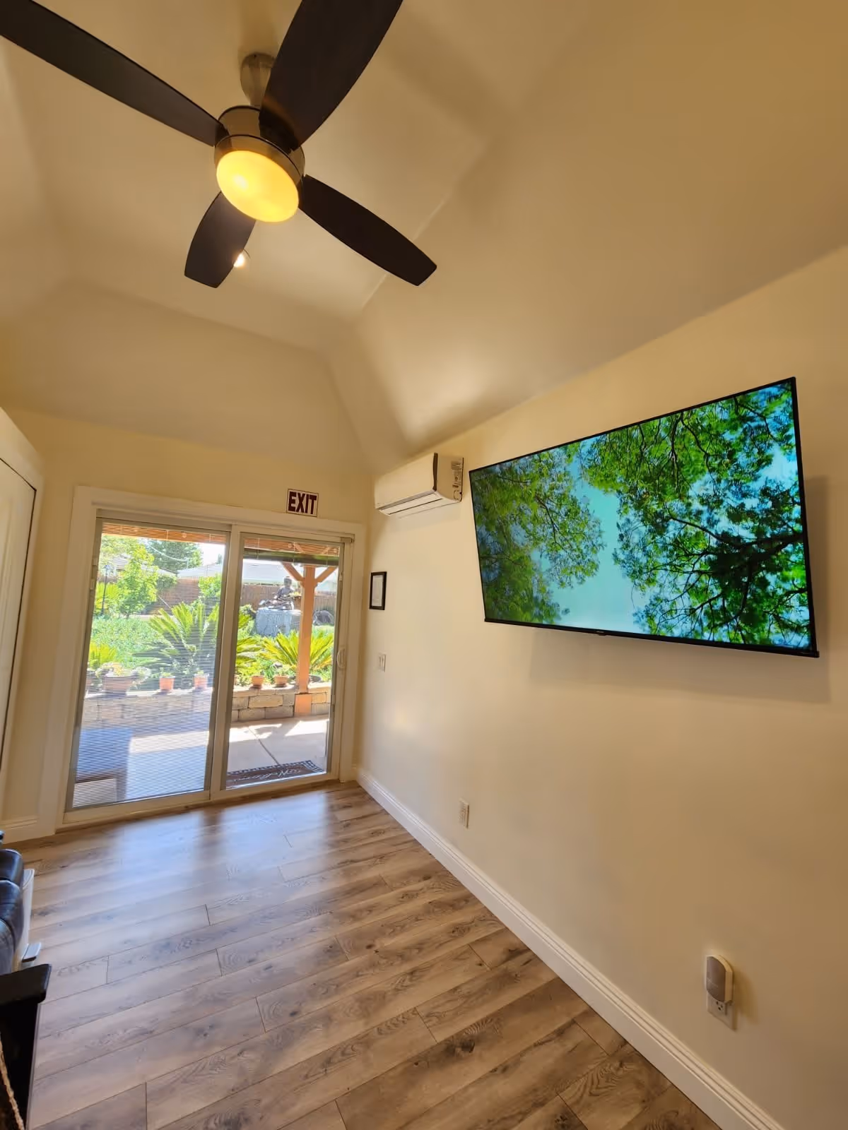 Interior room with wooden flooring, a ceiling fan with light, a wall-mounted flat screen TV displaying an image of tree branches and sky, a sliding glass door leading to an outdoor garden area, and an exit sign above the door.