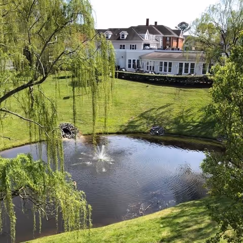 A serene outdoor scene featuring a small pond with a fountain in the center, surrounded by lush green grass and trees. In the background, there is a large white building with multiple windows and a sloped roof, partially obscured by the trees.