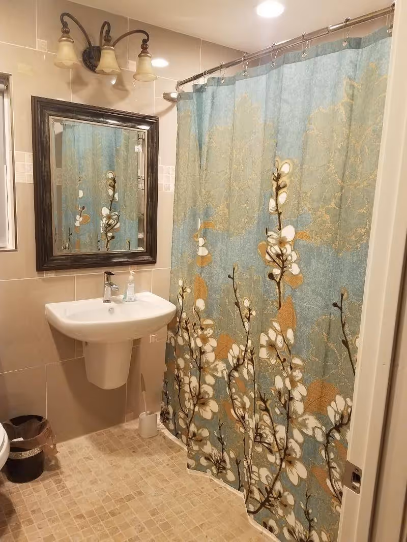 Bathroom with a pedestal sink, framed mirror, three-light wall sconce, floral shower curtain, and tiled floor.