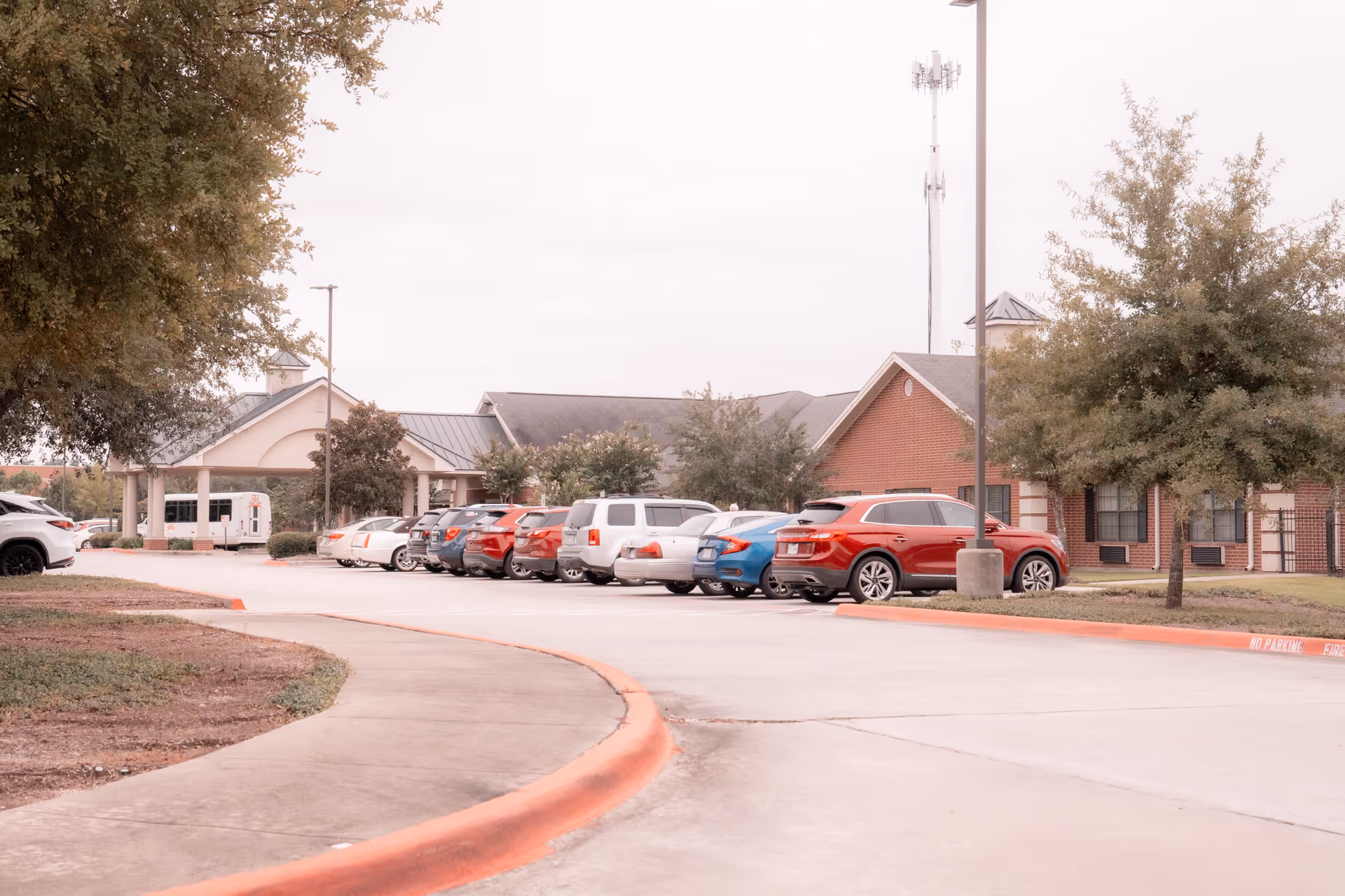 Front entrance and parking lot of a brick senior living facility with several parked cars and trees.