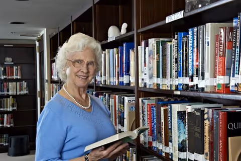 An elderly woman with white hair and glasses, wearing a blue sweater and a pearl necklace, is standing in a library holding an open book. She is smiling and looking at the camera, with shelves full of books behind her.