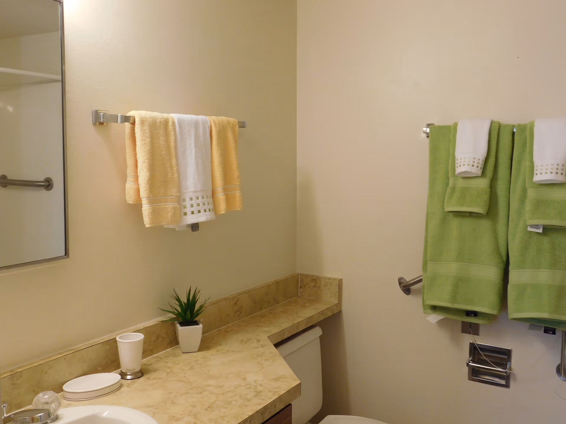 Bathroom interior with beige walls, a marble countertop with a small potted plant and a white cup, a sink, a mirror, and two towel racks holding yellow, white, and green towels.