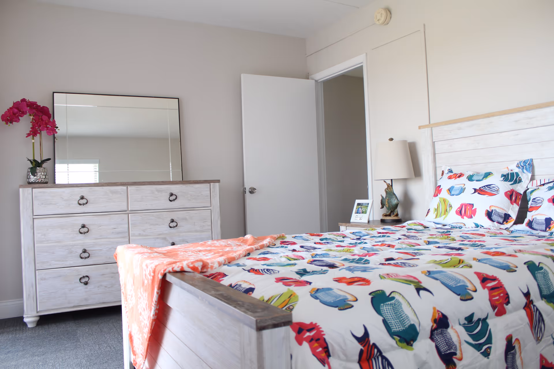 A bright bedroom with a white wooden bed featuring colorful fish-patterned bedding and matching pillows. A coral-colored blanket is draped over the foot of the bed. To the left, there is a white wooden dresser with a large mirror on top and a vase with pink flowers. A nightstand with a lamp and a framed photo is next to the bed. The walls are light-colored, and a door is open in the background.