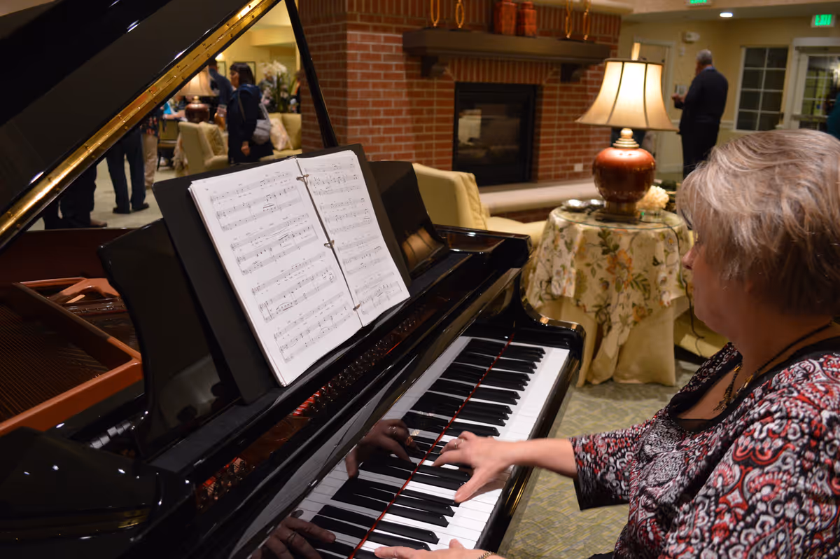 A person plays a grand piano in a communal lounge area with sheet music on the stand, a lamp-covered table, and a fireplace in the background.