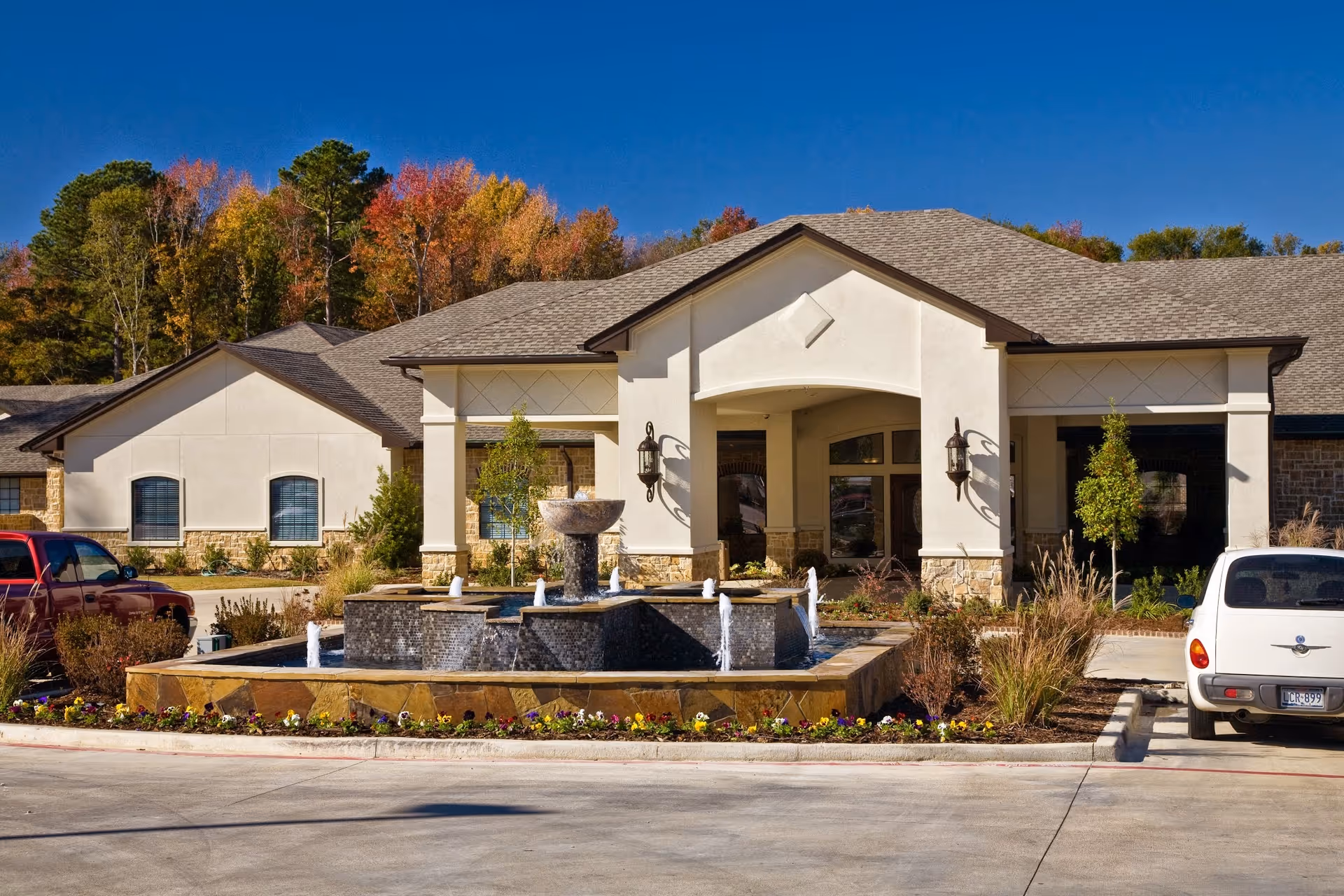 Front exterior view of Providence Park Rehabilitation And Skilled Nursing facility with a central water fountain, landscaped flower beds, and parked cars under a clear blue sky with autumn trees in the background.