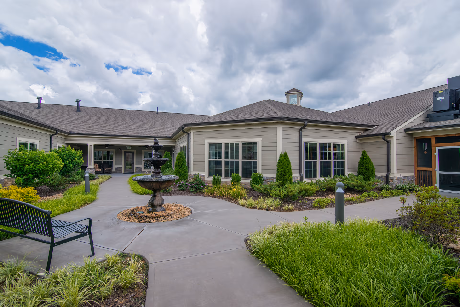 Outdoor courtyard area of a senior living facility with a central water fountain surrounded by landscaped greenery and bushes. There is a black metal bench on the left side and a paved walkway leading to the building entrance. The building has beige siding, multiple windows, and a cloudy sky overhead.