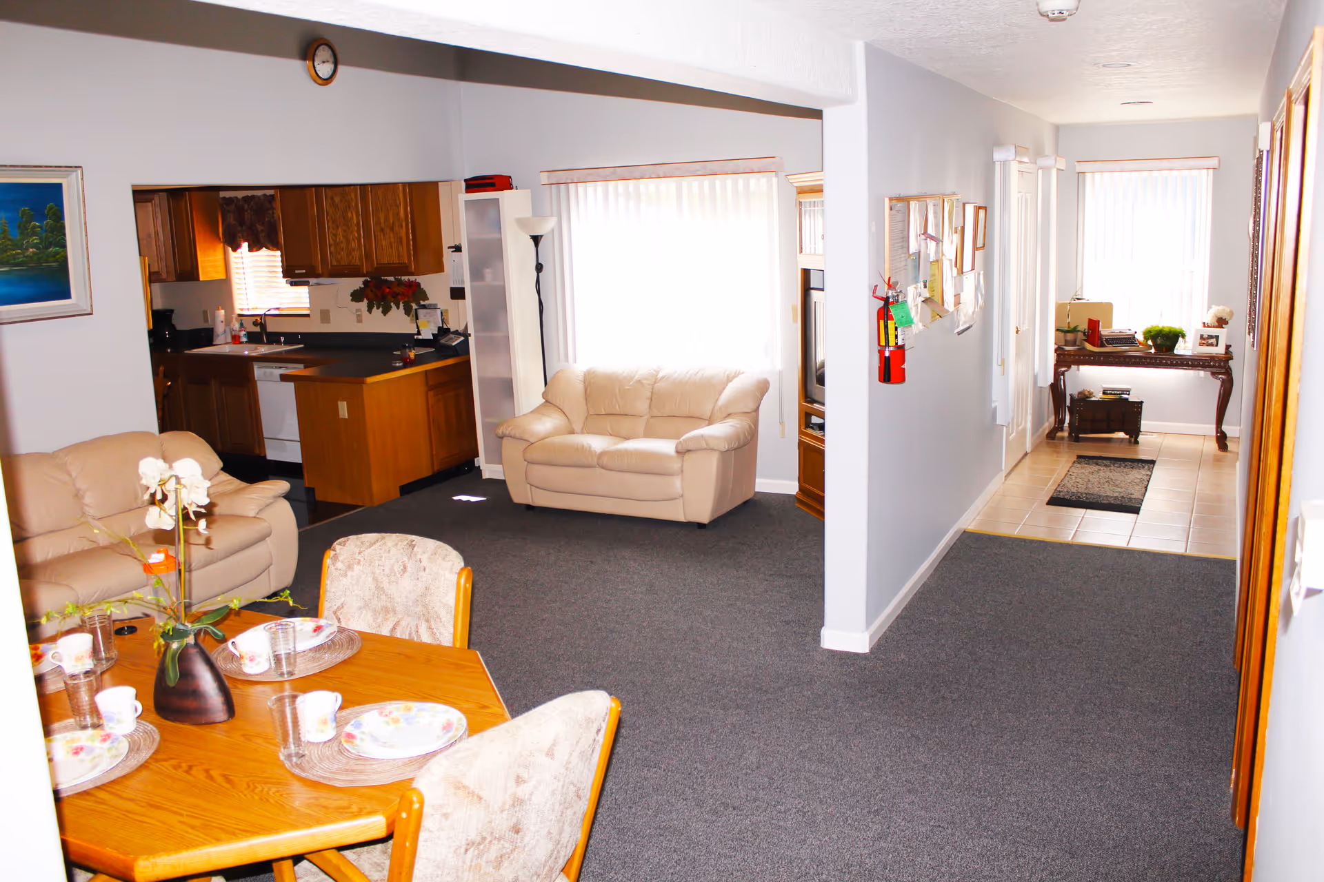 Interior view of a senior living facility showing a combined living and dining area. The living area has two beige leather sofas, a tall floor lamp, and a wooden TV cabinet. The dining area features a wooden table set with plates, cups, and glasses, surrounded by upholstered chairs. The kitchen with wooden cabinets and a dishwasher is visible in the background. A hallway with a bulletin board, fire extinguisher, and a small table with plants and decorations is seen to the right.