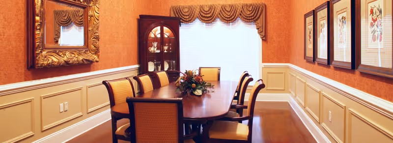Formal dining room featuring a long wooden table with upholstered chairs, a china cabinet, framed artwork, and a window with a valanced curtain.