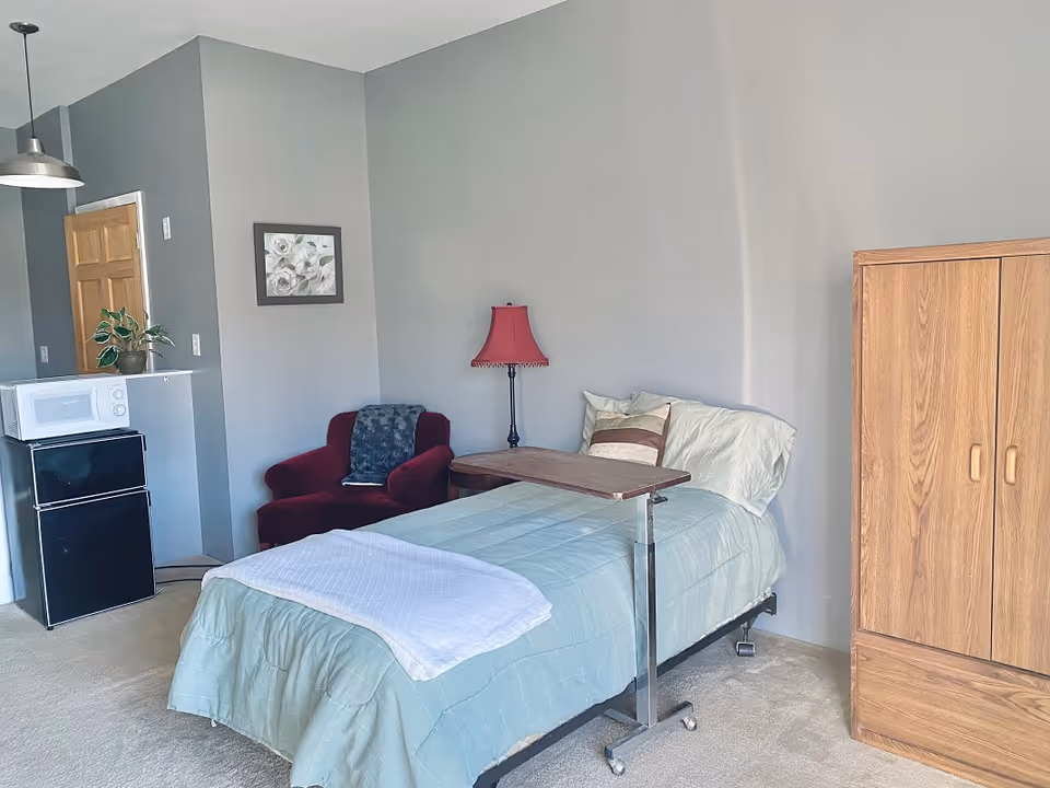 Single-occupancy bedroom with a bed and overbed table, red armchair, floor lamp, wooden wardrobe, and a small microwave on a mini-fridge.