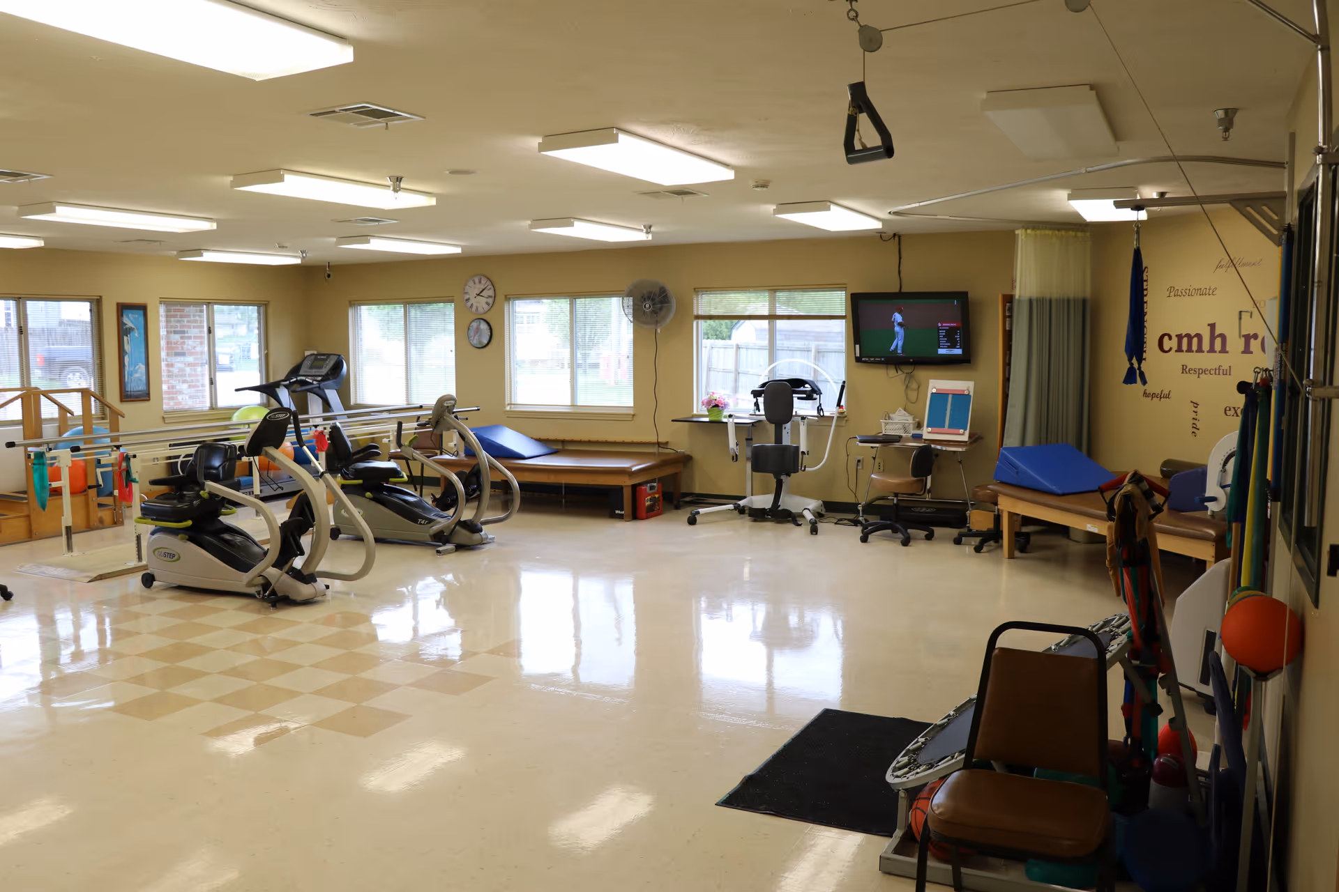 A spacious physical therapy or rehabilitation room with exercise equipment including stationary bikes, parallel bars for walking practice, therapy tables, chairs, and a wall-mounted TV showing a golf game. The room has large windows letting in natural light and motivational words on one wall.
