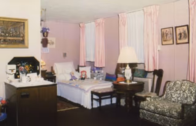 A cozy resident bedroom with a single bed, side table and lamp, patterned armchair, and pink curtains and walls.