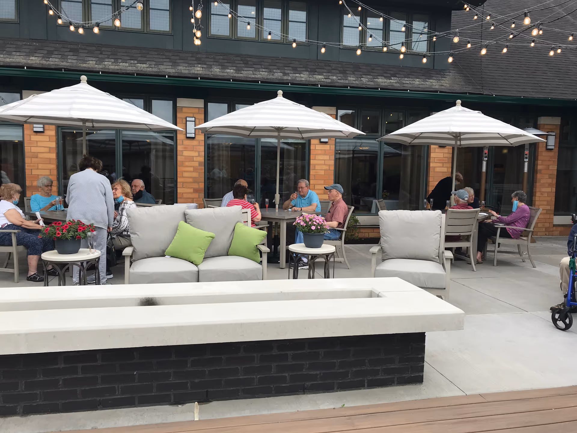 Outdoor patio area at The Summit at Brighton Independent Living with several elderly people sitting and socializing under large white umbrellas. The patio features cushioned seating with green pillows, small tables with flower pots, string lights overhead, and a modern fire pit in the foreground.