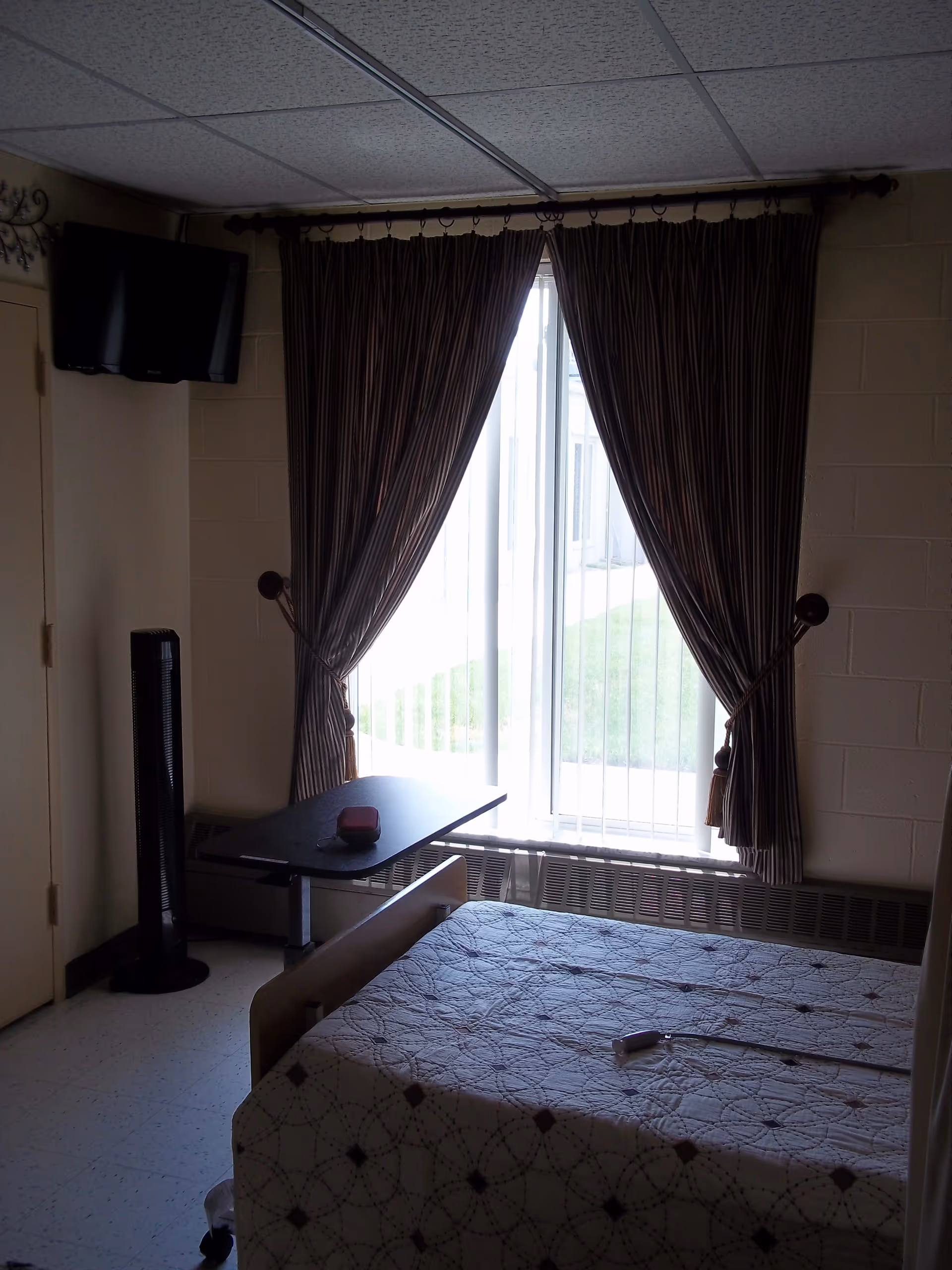A small bedroom in a senior living facility with a single bed covered by a patterned quilt. There is a window with vertical blinds and dark striped curtains tied back. A small black table is positioned near the window, and a tall black fan stands next to the wall. A flat-screen TV is mounted on the wall above the door.