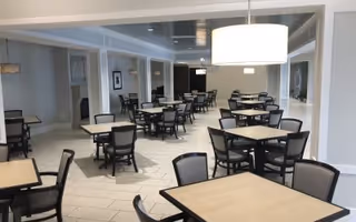 A spacious dining room with multiple square tables and black chairs arranged neatly. The room has light-colored tiled flooring, white walls, and large modern hanging light fixtures. There are windows and doorways visible along the walls.