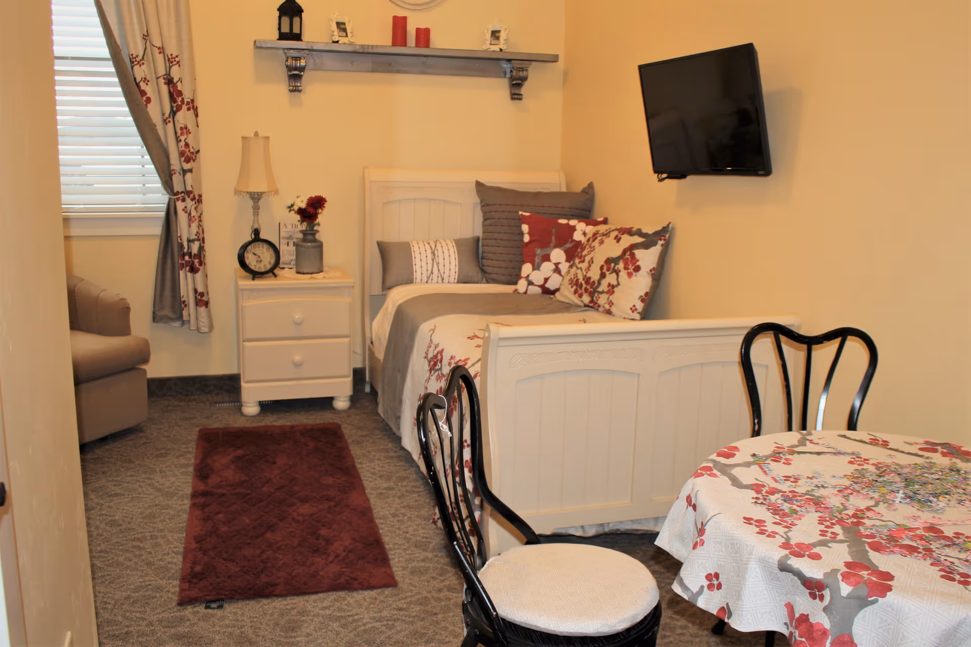 A cozy bedroom in an assisted living facility featuring a single bed with floral and striped pillows and a matching bedspread. Next to the bed is a white nightstand with a lamp, clock, and decorative items. A small round table with a floral tablecloth and two black chairs is in the foreground. A flat-screen TV is mounted on the wall, and a window with floral curtains is on the left side. The room has beige walls and carpeted flooring with a red rug.