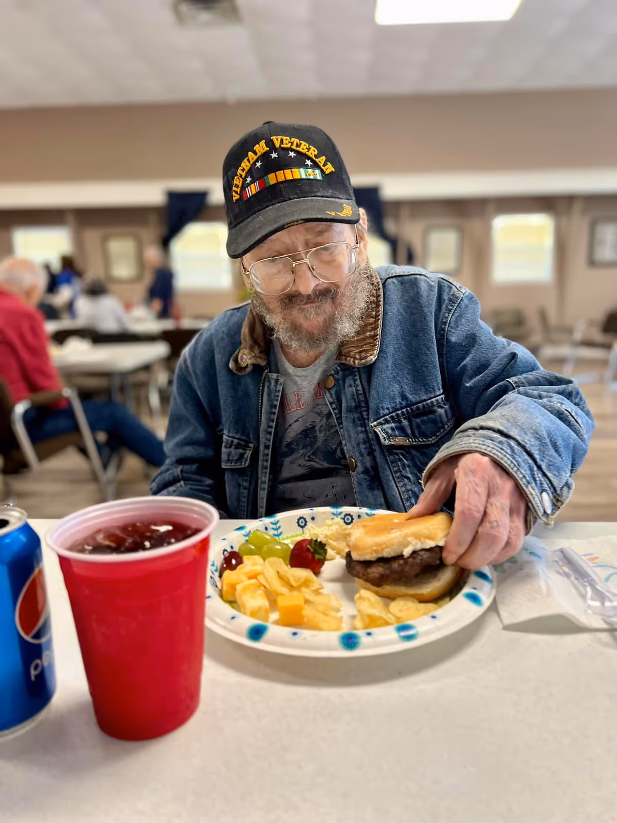 An older man in a 'Vietnam Veteran' cap and denim jacket sits in a dining hall holding a hamburger with a plate of chips, cheese and fruit and a red cup and soda can on the table.