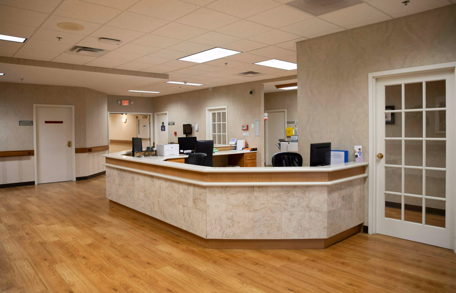 Reception desk and workstations in a well-lit senior living facility interior hallway with wood flooring and beige walls.