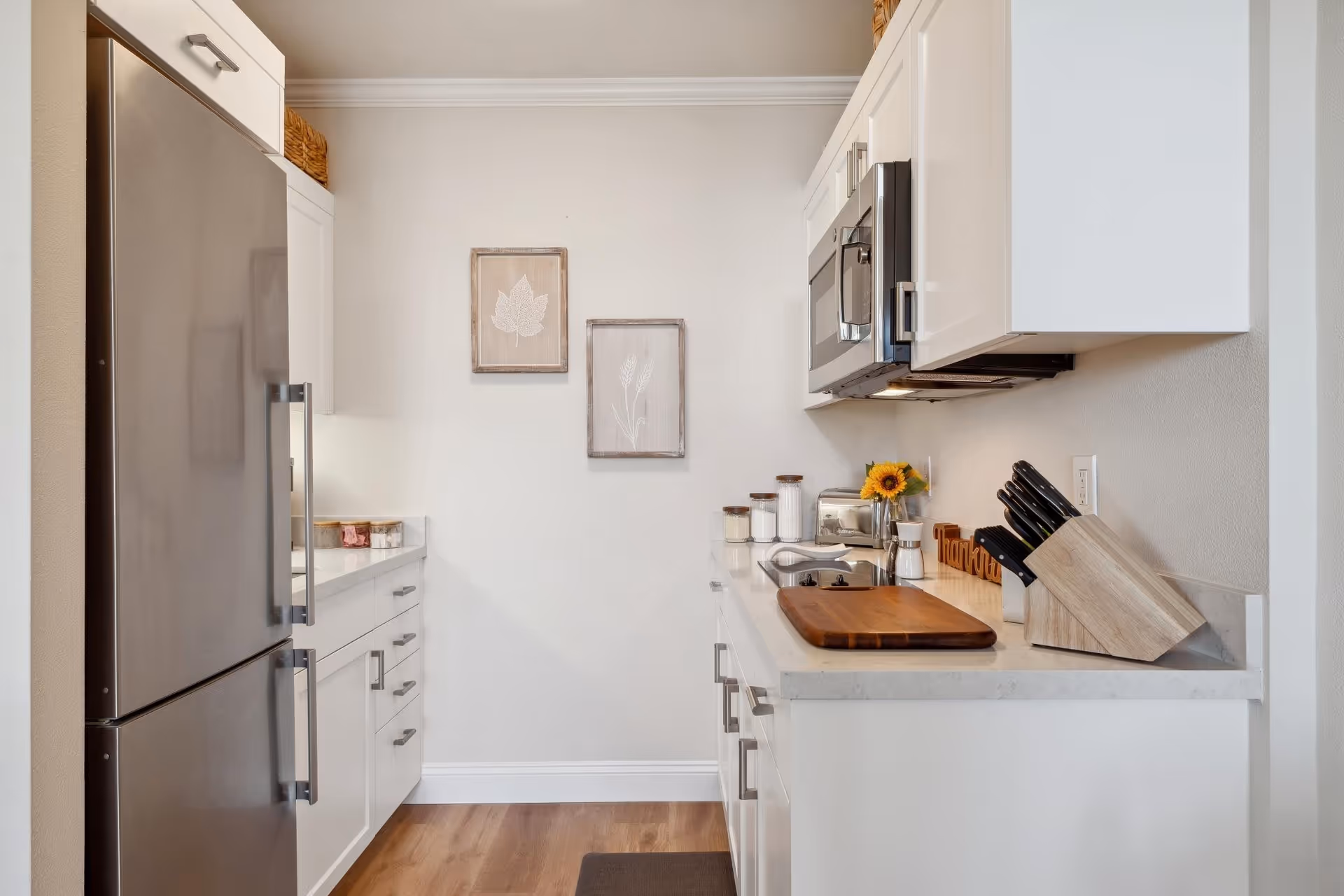 A modern kitchen with white cabinets, a stainless steel refrigerator, a microwave, a wooden cutting board on the countertop, a knife block, and decorative jars and flowers. Two framed botanical prints hang on the wall.