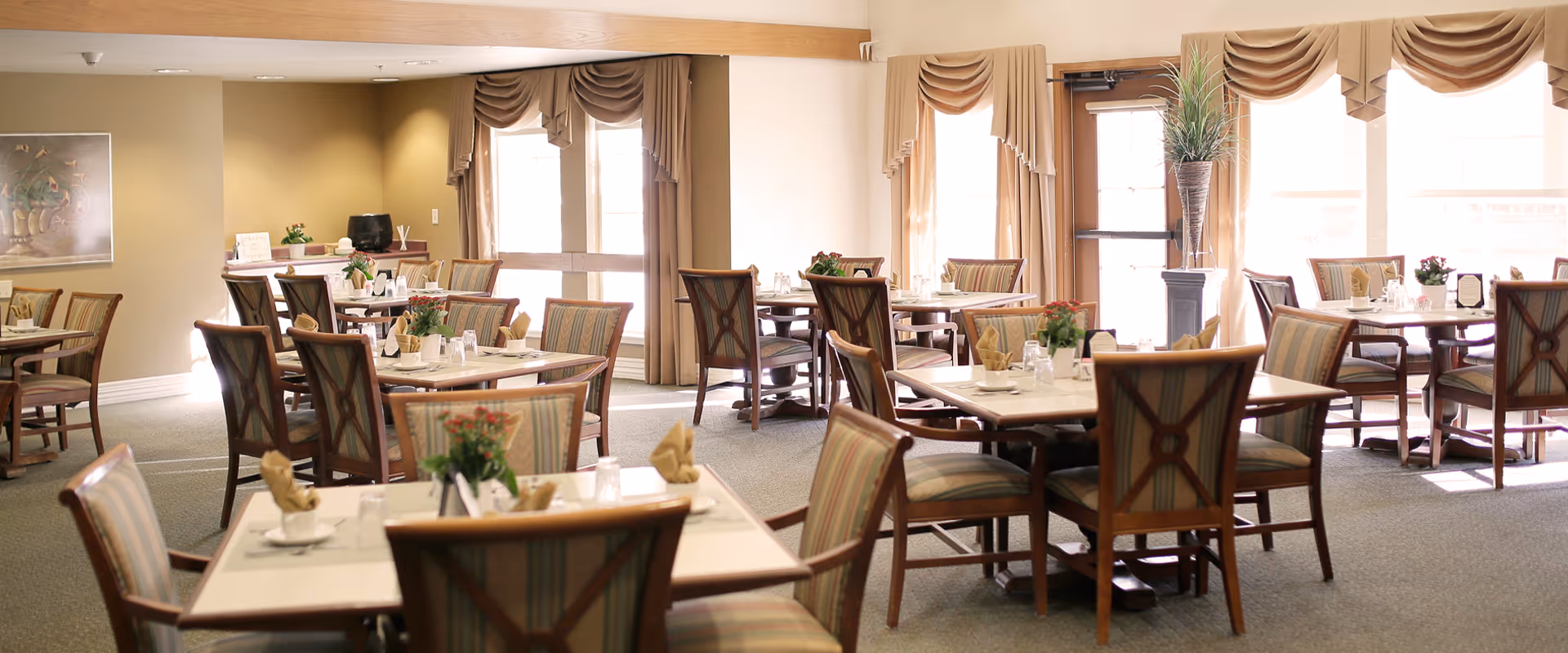 A bright dining room in a senior living facility with multiple square tables, each surrounded by four wooden chairs with striped cushions. The tables are set with napkins, glasses, and small flower arrangements. Large windows with beige curtains allow natural light to fill the room. The walls are painted a soft beige color, and there is a framed painting on one wall.