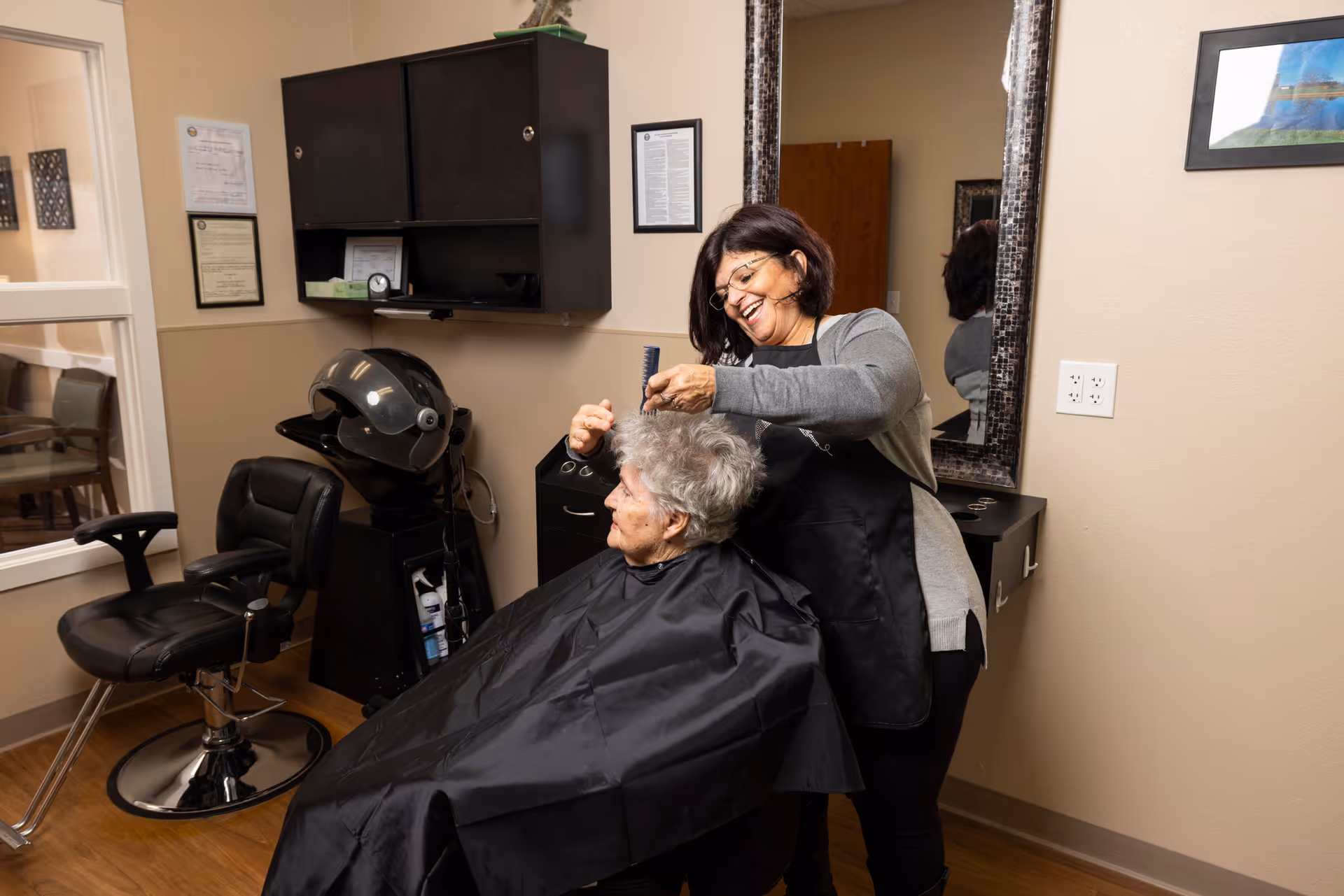 A hairstylist cutting an elderly woman's hair in a salon chair inside a senior care facility.
