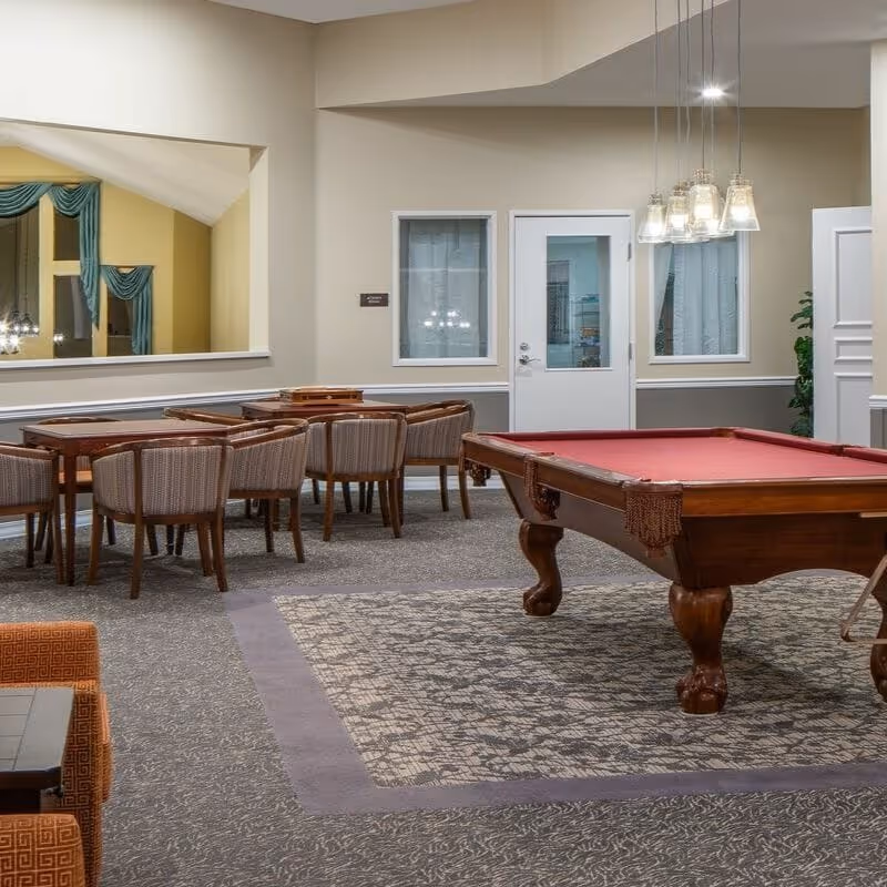 A senior living facility common area featuring a pool table with a red felt top, several wooden tables surrounded by cushioned chairs, a large mirror on the wall, and pendant lights hanging from the ceiling. The room has neutral-colored walls and carpeted flooring with a patterned rug under the pool table.