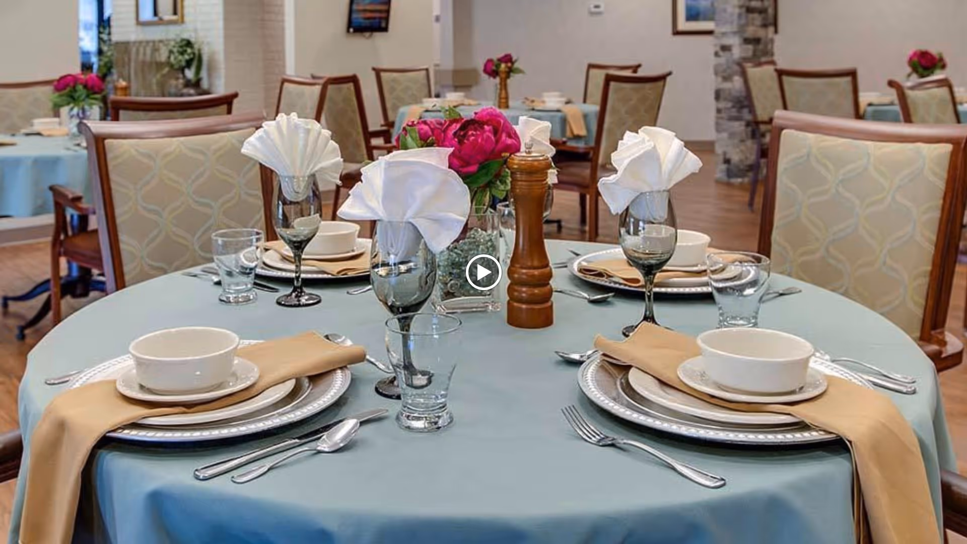 A round dining table set for four with light blue tablecloth, beige napkins, white plates, bowls, silverware, water glasses, wine glasses with folded white napkins, and a centerpiece of pink flowers and a wooden pepper grinder in a senior living facility dining room.