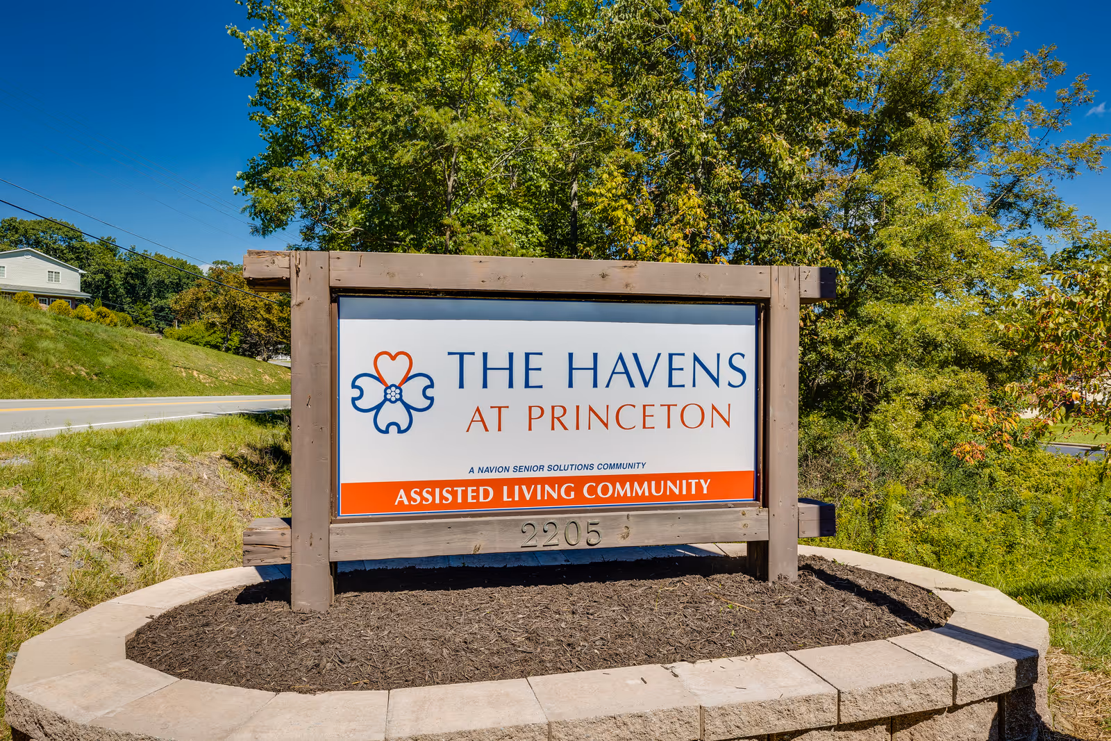 Outdoor wooden sign for The Havens at Princeton, an assisted living community, surrounded by greenery and trees under a clear blue sky.