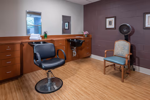 Salon-style room with a black stylist chair, wash basin, hooded hair dryer, and an upholstered waiting chair beside wood cabinets and a painted cinderblock wall.