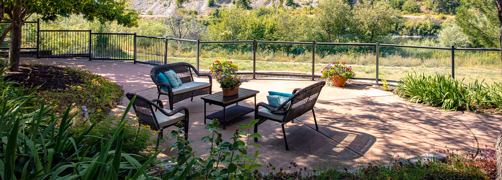 Outdoor paved patio with wicker chairs, a coffee table, potted plants, and a railing overlooking greenery.