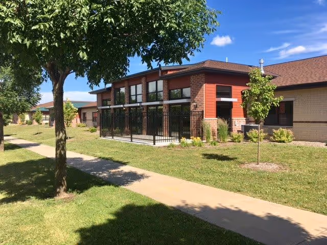 Exterior view of a single-story senior living facility building with a brick and siding facade, large windows, and a fenced patio area. The building is surrounded by a well-maintained lawn, trees, and a concrete walkway under a clear blue sky.