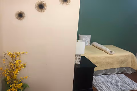 A neatly decorated bedroom with a single bed against a green accent wall, a nightstand with a lamp, wall art, and a floral arrangement.