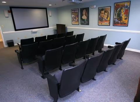 Small movie theater room with three rows of black theater seats facing a large projection screen on a light blue wall. Four vintage movie posters are displayed on the right wall.