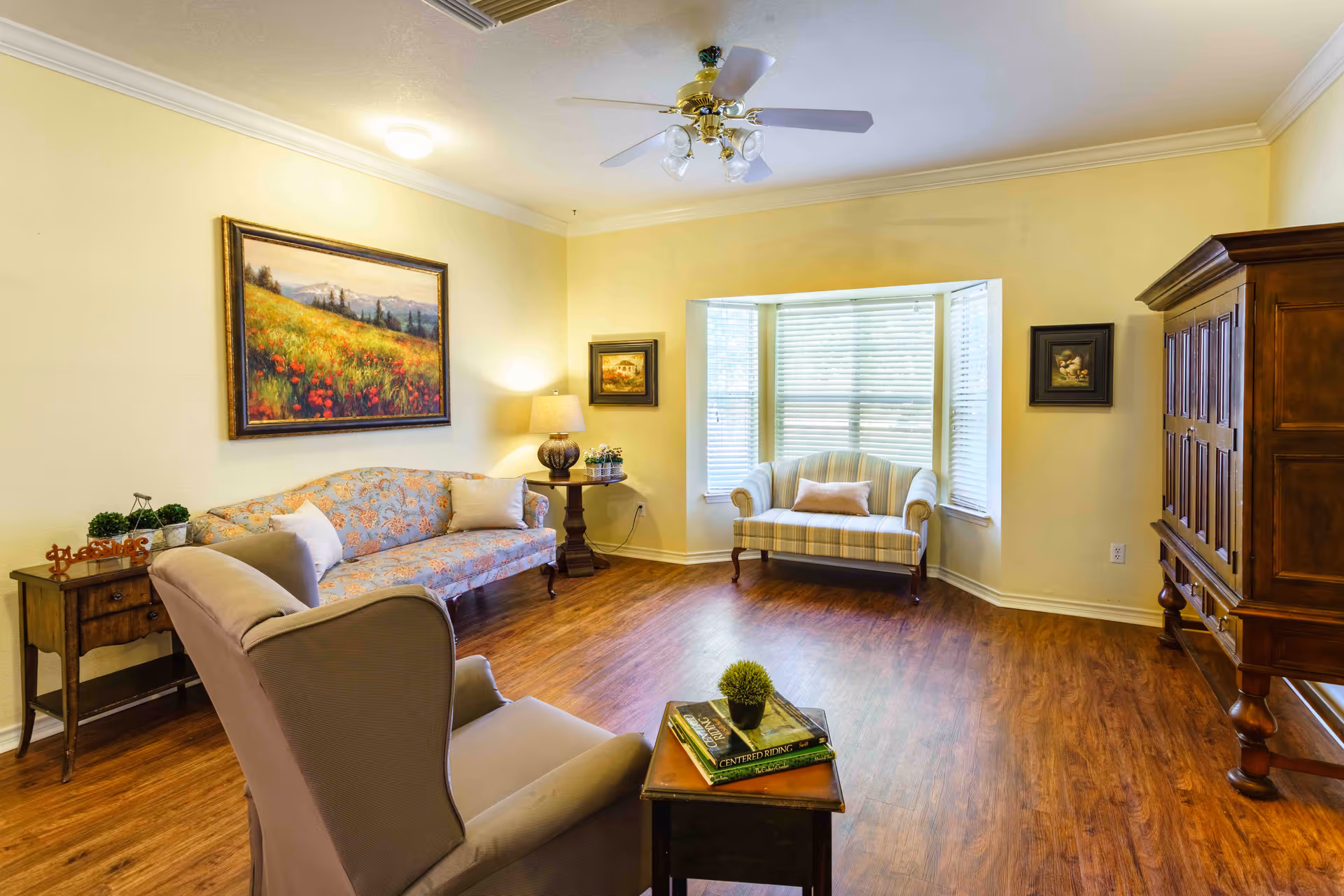 A cozy living room with wooden flooring, featuring a floral patterned sofa, a striped loveseat by a bay window with blinds, a cushioned armchair, a wooden side table with books and a small plant, a wooden cabinet, and framed paintings on the yellow walls. A ceiling fan with lights is mounted on the ceiling.