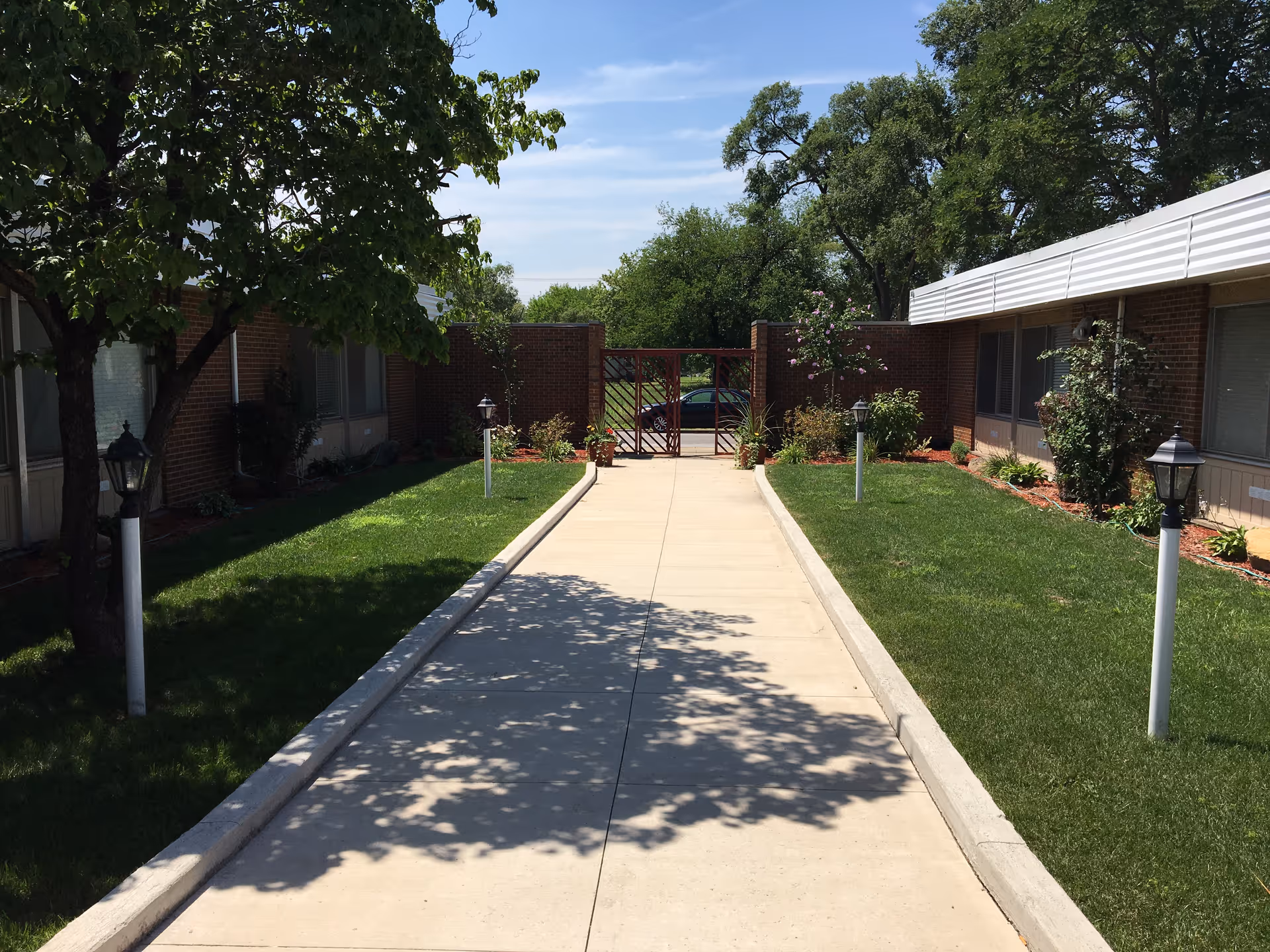 A paved walkway flanked by green lawns and small garden beds with shrubs and trees, leading to a closed gate. The walkway is bordered by lamp posts on both sides, and the area is surrounded by single-story brick buildings under a clear blue sky.