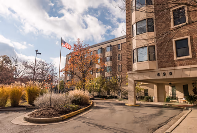Front entrance of a brick senior living building with address 690, a circular driveway, American flag, and autumn landscaping.