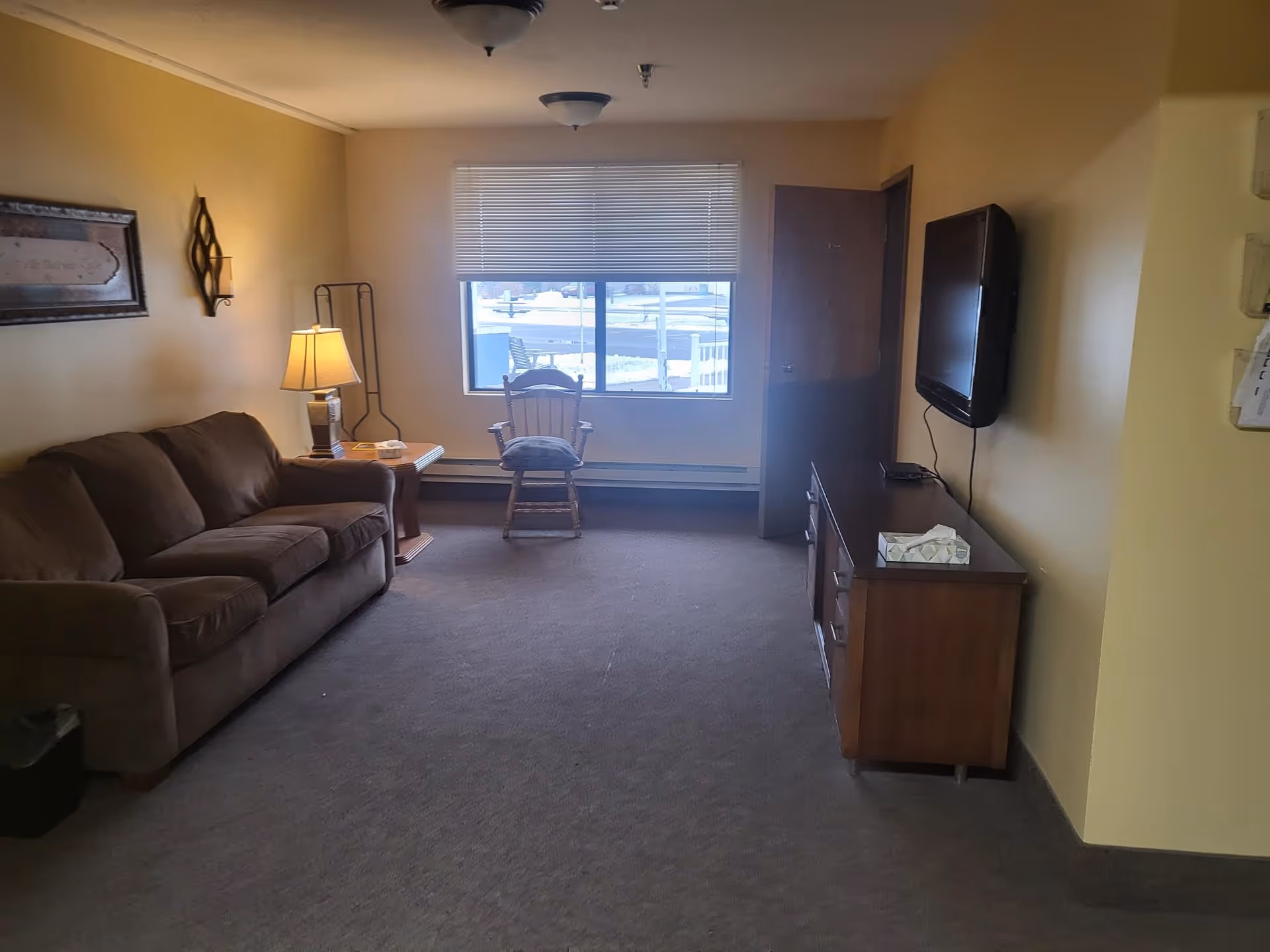 A cozy living room with a brown sofa on the left, a wooden rocking chair near a window with blinds, a wooden side table with a lamp, and a wooden TV stand with a flat-screen TV mounted on the wall. The walls are painted light yellow and the floor is carpeted.