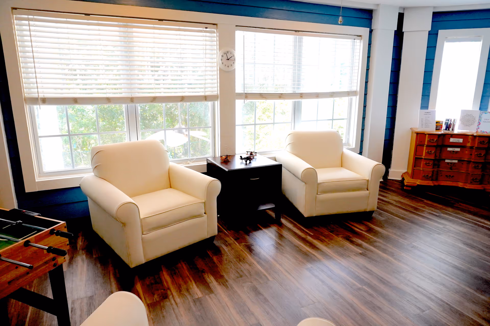 A bright room with two white armchairs positioned around a small dark wooden table. Behind the chairs are large windows with white blinds partially open, letting in natural light. A clock is mounted on the wall between the windows. To the right, there is a wooden chest of drawers with various items on top. The floor is dark wood, and part of a foosball table is visible on the left side of the image.