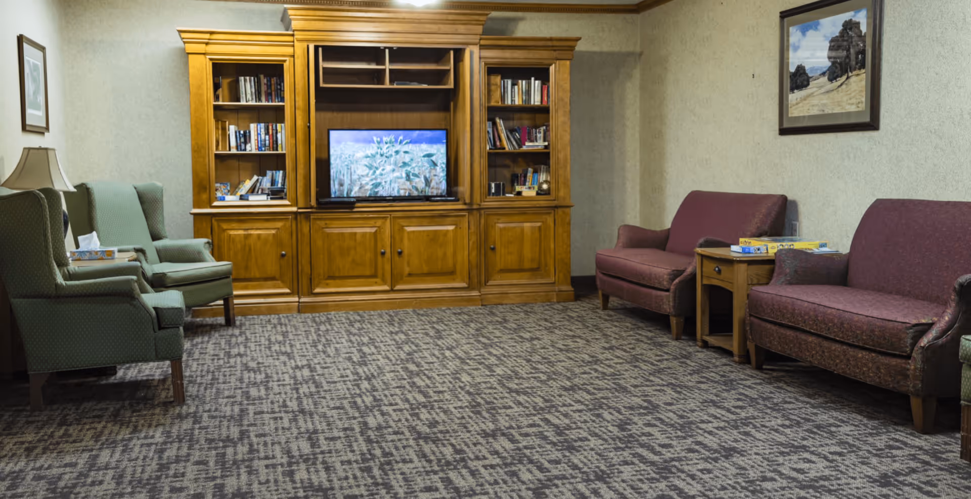 A cozy living room area with two green armchairs on the left and two maroon sofas on the right, separated by a wooden side table with board games on it. A large wooden entertainment center with shelves filled with books and a TV is centered against the back wall. The room has patterned carpet and framed artwork on the walls.