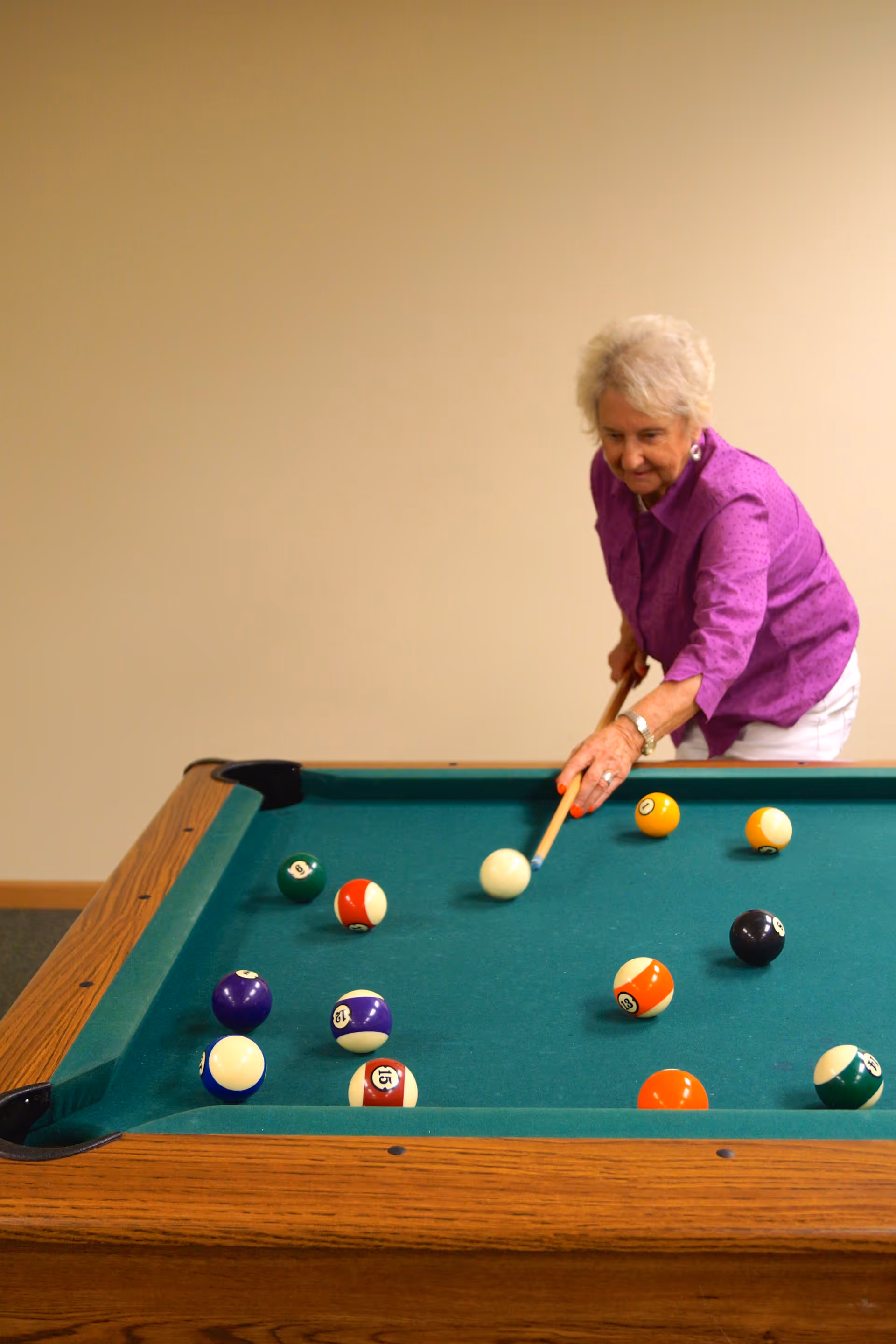 An elderly woman wearing a purple shirt and white pants is playing pool, aiming a cue stick at the white cue ball on a green felt pool table with several colored balls scattered across it.