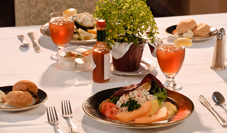 Table set with plated salad, bread rolls, iced tea glasses, a bottle of hot sauce, and a potted plant on a white tablecloth.