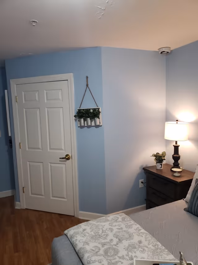 A cozy bedroom corner with light blue walls, a white door, and a wooden floor. There is a small dark wooden nightstand with a lamp and a small plant on it next to a bed with a gray quilt and a patterned blanket. A decorative hanging planter with green plants is mounted on the wall.