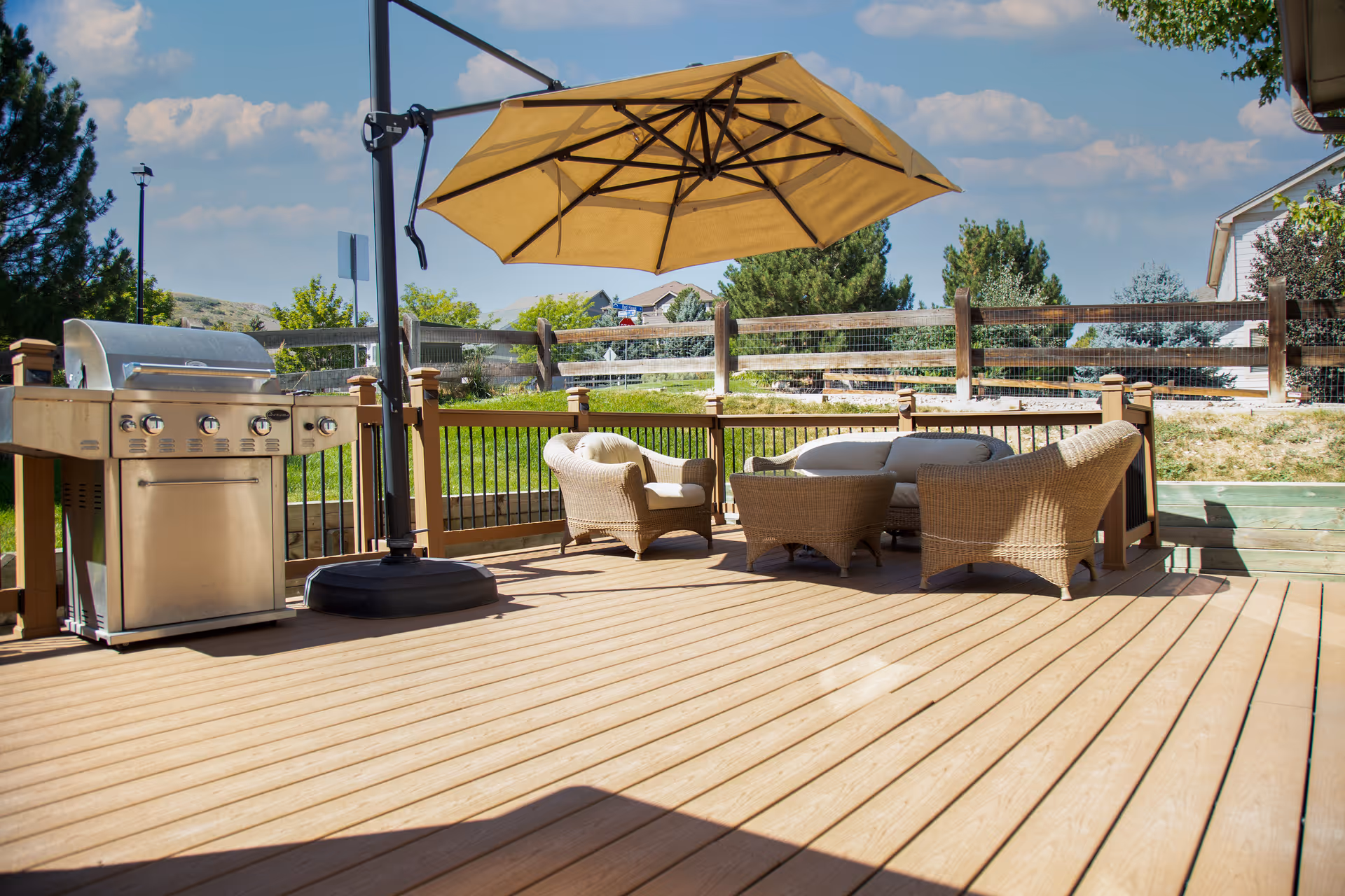 Sunny outdoor wooden deck with a BBQ grill, wicker seating under a large yellow umbrella and a view of a fenced yard.