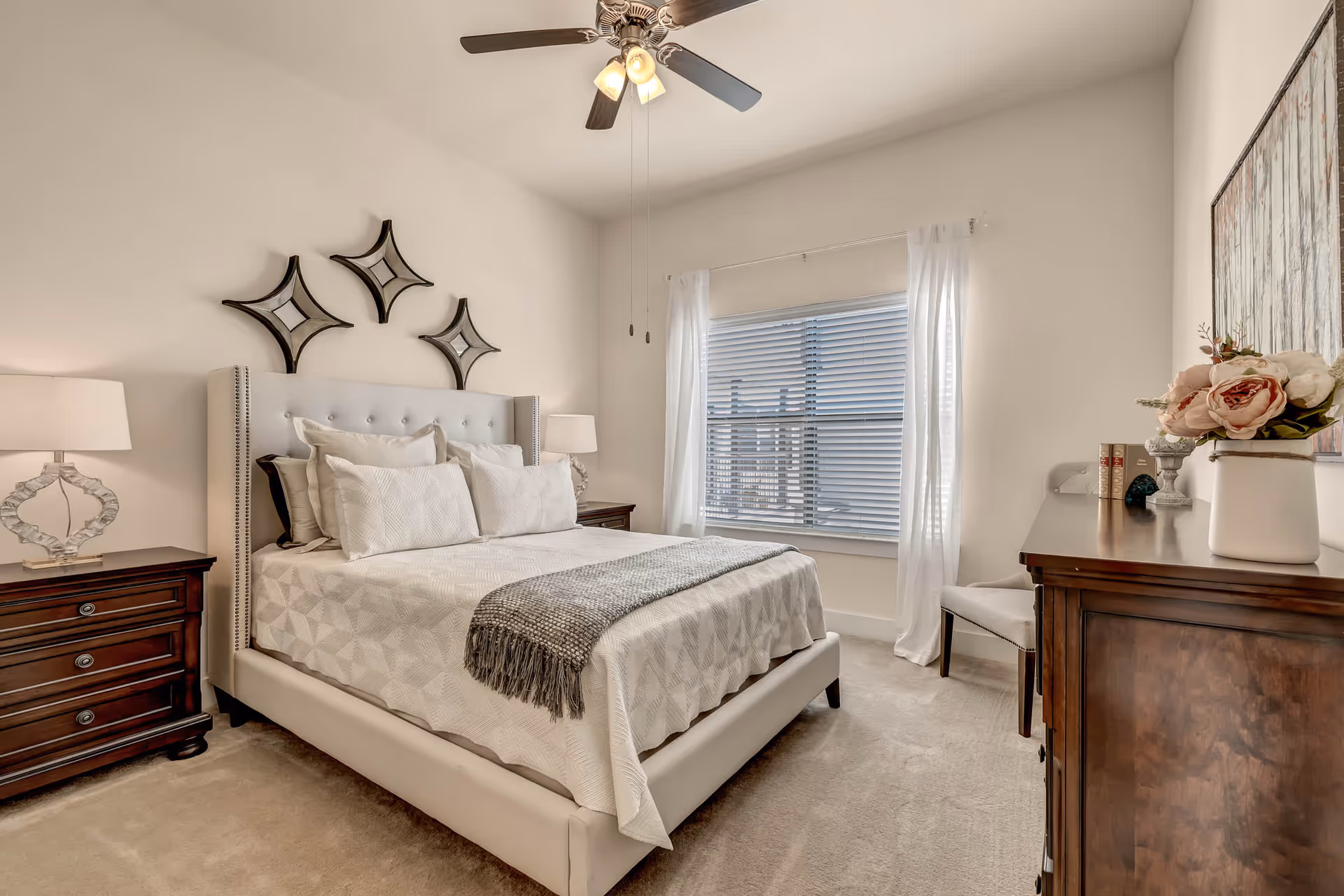 A neatly arranged bedroom with a large bed featuring white bedding and multiple pillows. There are two dark wooden nightstands with modern lamps on either side of the bed. Above the bed, there are three decorative star-shaped mirrors on the wall. A window with white curtains and blinds lets in natural light. A wooden dresser with a vase of flowers and some books is positioned on the right side of the room, along with a white chair. The ceiling has a fan with lights.