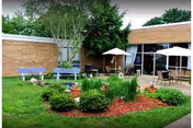 Outdoor garden area with green grass, bushes, and small trees in front of a brick building. There are patio tables with umbrellas and chairs on a paved area near the building.