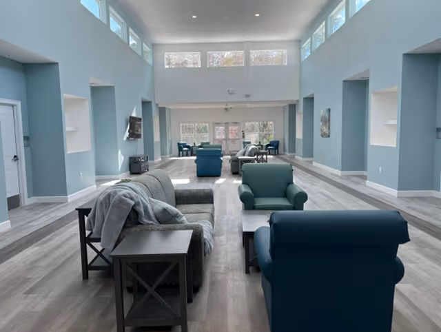A spacious senior living facility common area with light blue walls and large windows near the ceiling allowing natural light to fill the room. The room is furnished with multiple armchairs and sofas arranged around small tables on a light wood floor. A flat-screen TV is mounted on the left wall.
