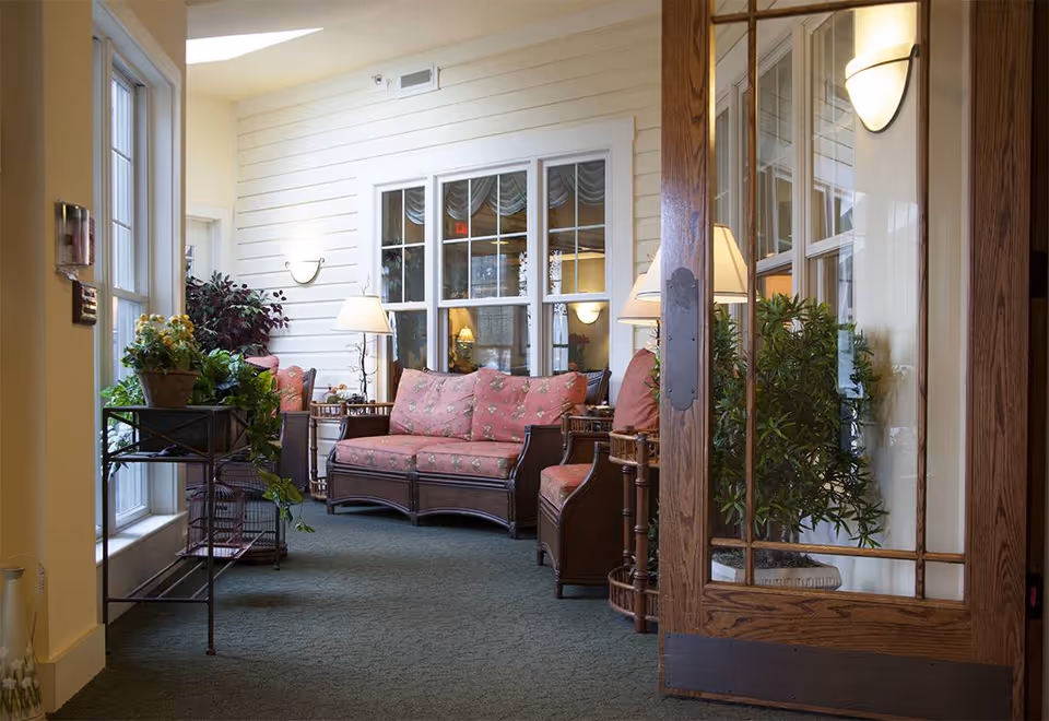 A cozy indoor sitting area with floral cushioned wicker furniture including a sofa and chairs, several potted plants, a floor lamp, and a wooden framed glass door partially open, with large windows letting in natural light.