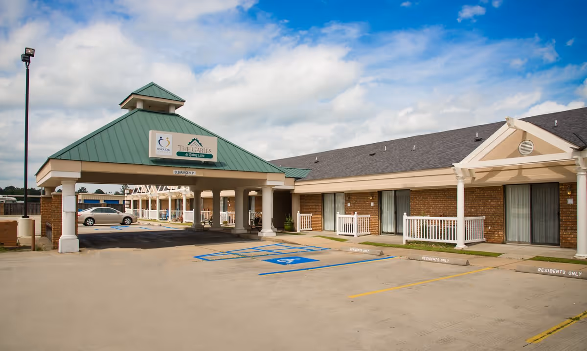 Exterior view of The Gables At Spring Lake Assisted Living facility showing a covered entrance with a green roof and columns, adjacent to a parking area with designated handicapped and residents-only parking spaces. The building has brick walls and sliding glass doors with small white fences in front of each unit.