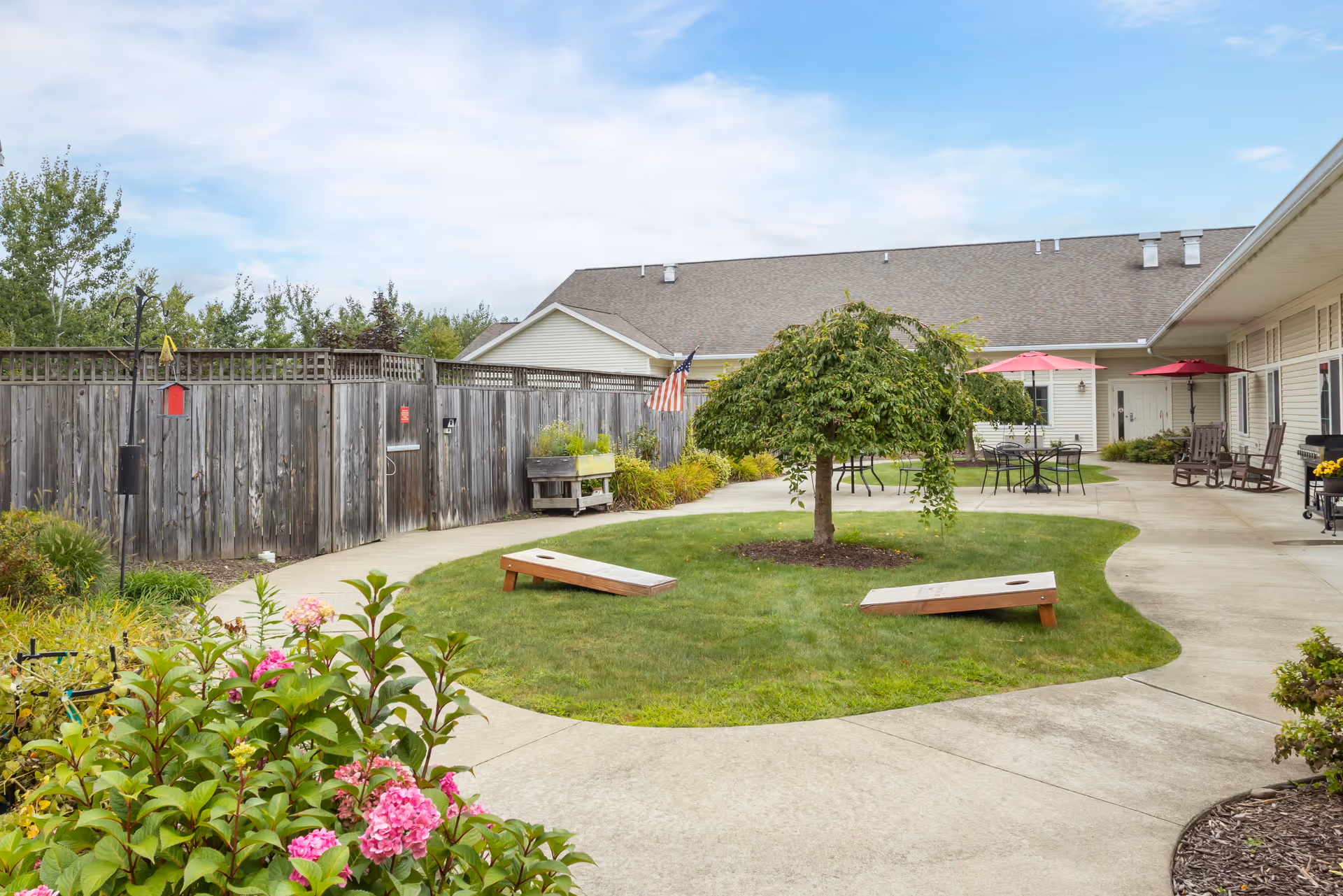 Outdoor courtyard area with a small tree in the center of a grassy patch surrounded by a concrete walkway. Two wooden cornhole boards are placed on the grass. There are patio tables with umbrellas and chairs along the walkway near the building. A wooden fence encloses the area, and some flowering plants are visible in the foreground.