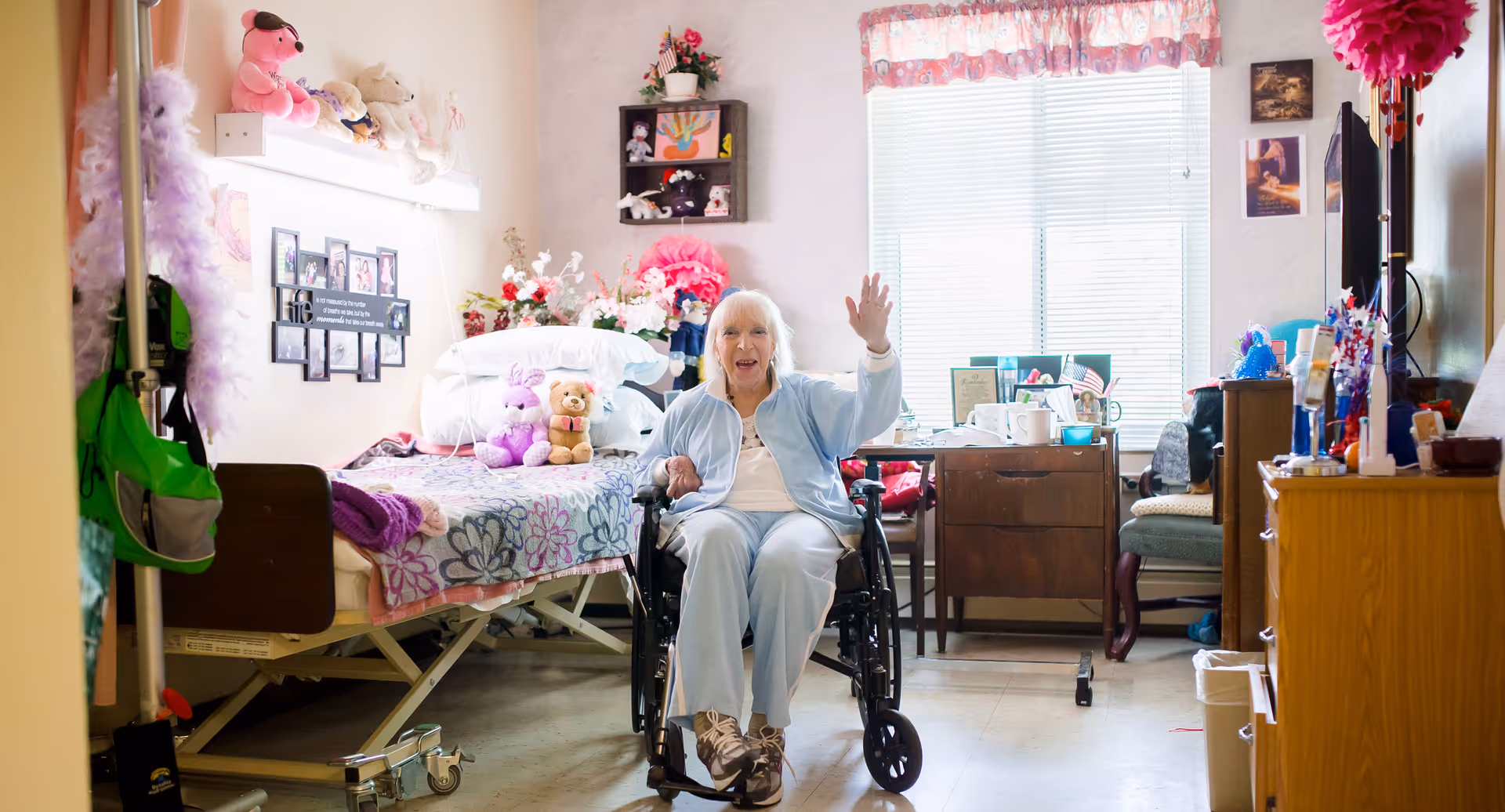 An elderly woman in a wheelchair waving inside a decorated bedroom with a bed, stuffed animals, flowers, and various personal items. The room has a window with blinds and floral curtains, a wooden dresser, and a chair.