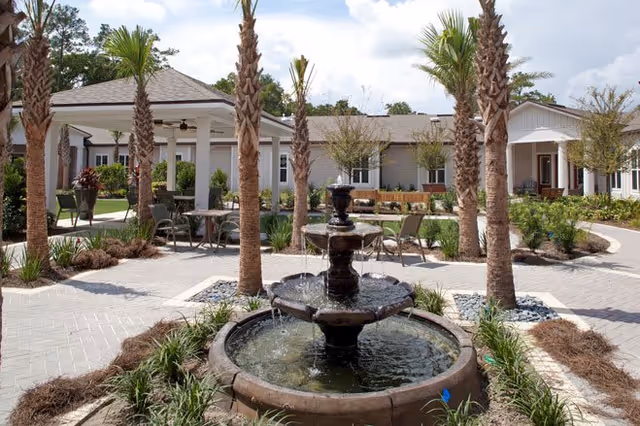 Outdoor courtyard area at Ortega Gardens featuring a central three-tiered water fountain surrounded by palm trees, paved walkways, garden beds with plants, and seating areas with tables and chairs under a covered gazebo and open sky.